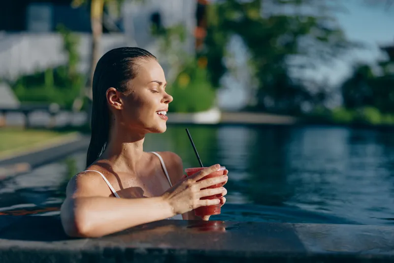 Cheerful woman in a pool, holding a red beverage, sunlight on skin, relaxed smile, leaning on the pool edge, summer vibe, outdoor leisure, vibrant colors, calm moment by water, carefree lifestyle,