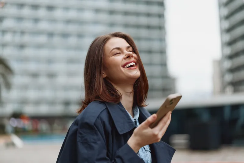 Happy woman outdoors smiling with eyes closed holding smartphone urban background casual clothing natural light positive emotion