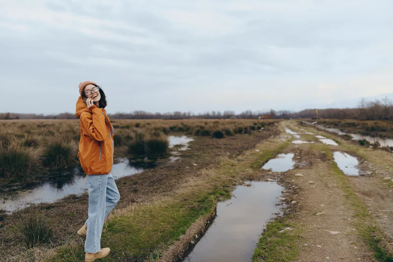 Woman lifestyle outdoors in rainy cloudy weather, standing beside puddles on a muddy path in countryside nature, wearing coat and jeans, calm travel portrait near wet grass