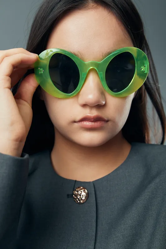Young woman with long dark hair wearing large green sunglasses and a dark jacket with a gold brooch posing against a plain background for fashion portrait.