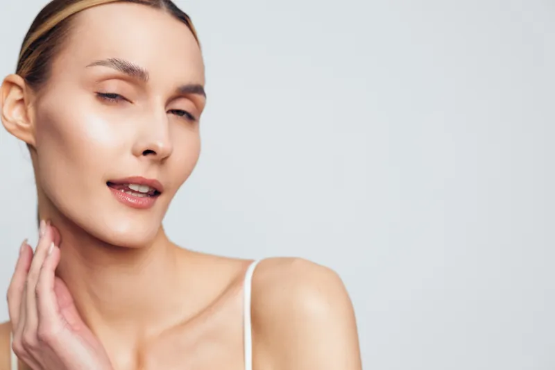 Smiling woman with smooth skin applying skincare routine. Close-up on a light background. Concept of beauty, skincare, and selfcare. Soft studio lighting creates a calm, confident mood.