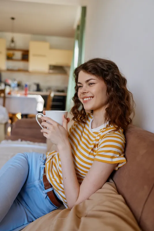 Smiling woman relaxing at home, sitting on a sofa with a mug in hand, wearing a striped shirt, cozy living room setting, casual lifestyle moment indoors.