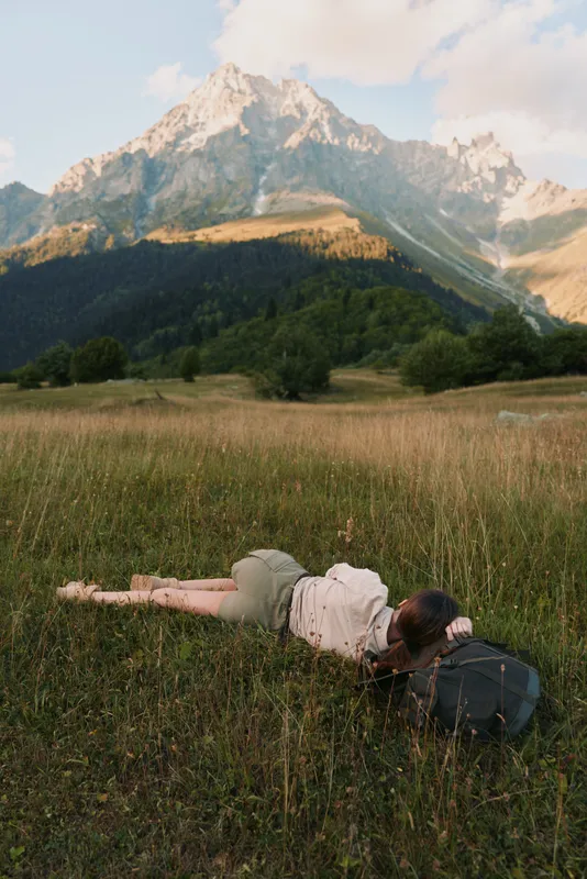 Young woman with long hair lies on grass in a mountain meadow resting her head on a backpack and enjoying nature during a peaceful summer day.