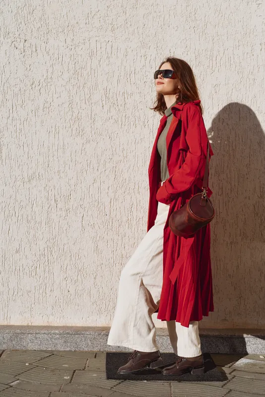 Young woman in a red trench coat walks confidently along a sunlit street wearing sunglasses and white pants with a brown leather handbag.
