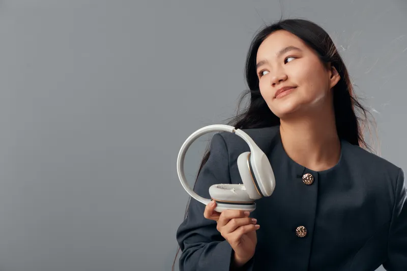 Young woman in dark jacket holding white headphones looking sideways with slight smile against gray background. Female adult posing with wireless audio device in casual style.