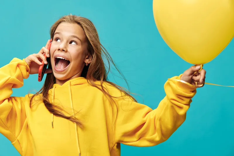 Girl in yellow hoodie talking on phone and holding yellow balloon with happy expression and open mouth on blue background for celebration and communication.