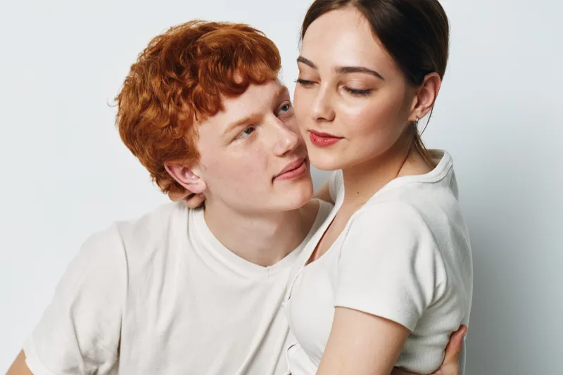 Couple embracing with affection, young man and woman in casual white clothes posing on light background, closeup portrait showing intimacy and connection between partners.