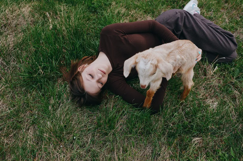 Young woman with long brown hair lies on green grass smiling gently while a small baby goat stands nearby in a relaxed outdoor lifestyle scene during daytime.
