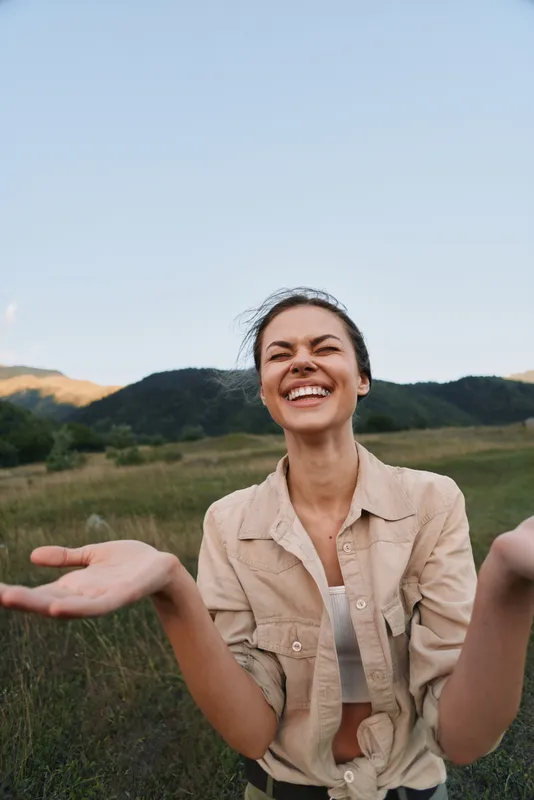 A happy young woman in casual clothes smiles broadly with open arms in a green field with hills in the background during daylight. Concept for lifestyle and joy.