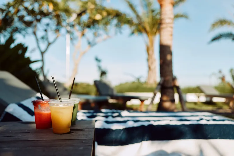 Cheerful poolside scene with two colorful beverages on a wooden table, tropical palms in the background, sunlit lounge, striped towels, relaxed lifestyle moment, vibrant colors, fresh summer vibe