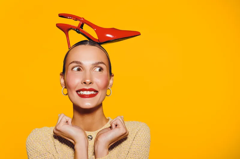 Young woman with red high heel shoe on her head smiles playfully against bright yellow background. Concept of fashion, fun, creativity, and joyful expression in studio lighting.