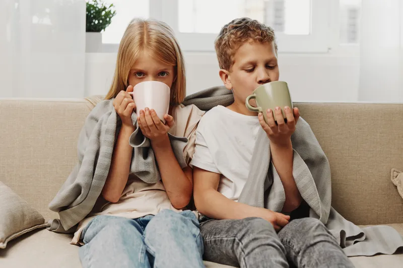 Two children wrapped in a cozy blanket sit on a sofa drinking hot beverages from mugs in a bright living room. Concept for warmth, comfort, family moments, and relaxing indoors.