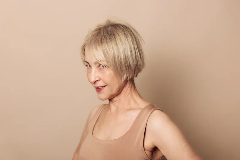 Confident mature woman with short blonde hair wearing a sleeveless beige top poses against a plain beige background. Concept of natural aging, self-assurance, and minimalistic portrait.
