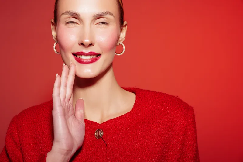 Smiling woman in red sweater waves hand on red background. Close-up portrait with bright lipstick and hoop earrings. Concept of greeting, fashion, and positive emotions in studio lighting.