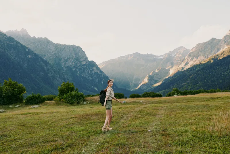 Young woman hiking in mountain valley, wearing backpack and shorts, enjoying nature, sunny day, green grass, rocky peaks in background, active lifestyle outdoors