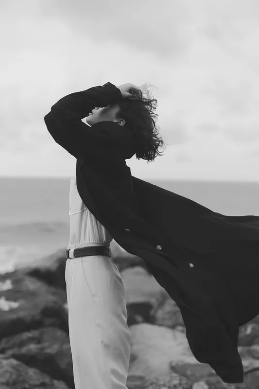 Cape waves with the wind against a vast seascape, a figure stands on rocky shore, white attire contrasted by dark cape, atmosphere bold and cinematic, conveying motion, freedom and dramatic style