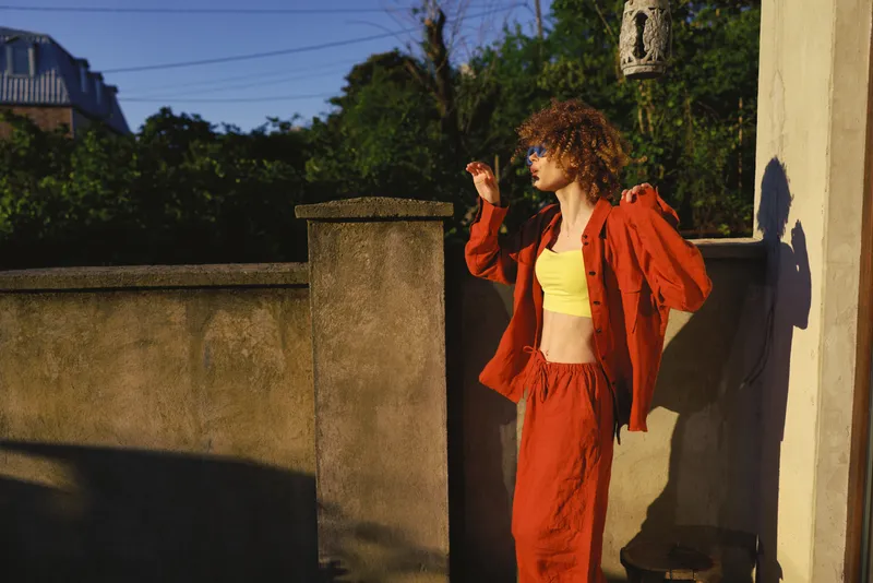 Young woman in a bright red tracksuit leaning against a concrete wall, yellow crop top, basking in sunlight. Outdoor lifestyle, casual fashion, confidence, and relaxed mood