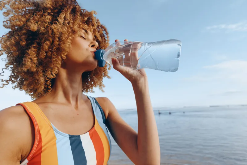 Woman, curly hair, drinking water, hydration, ocean, outdoors, summer, travel, backpack, seaside, young female sips cool water from bottle by blue horizon in bright sunlight