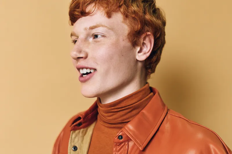 Young man with red hair wearing orange turtleneck and leather jacket smiling looking aside on beige background in studio portrait with natural emotion and casual style.