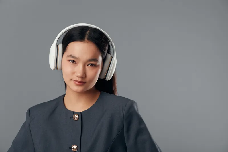 Young woman wearing wireless headphones and dark jacket standing against gray background, looking at camera with confident expression and relaxed posture in studio.