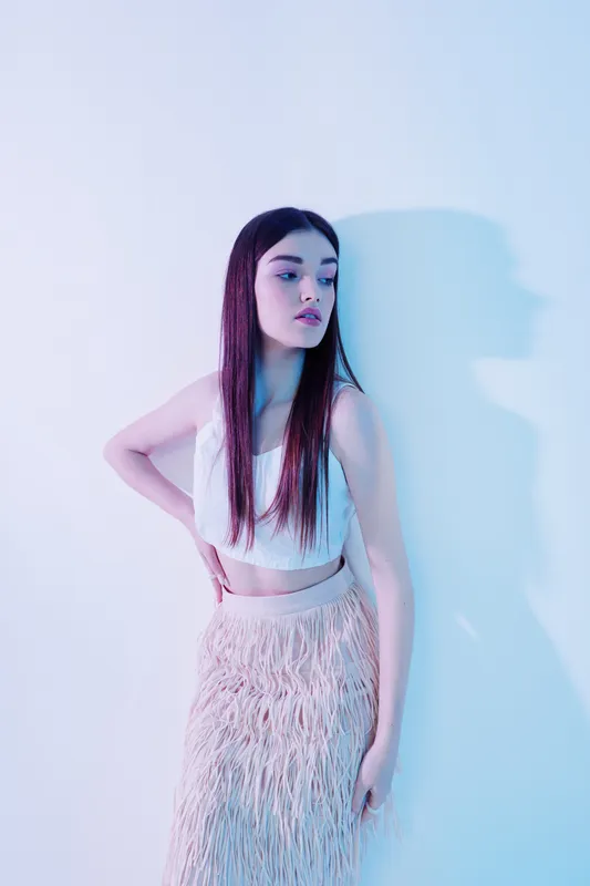 Young woman beauty fashion model poses in a white crop top and textured skirt against a light background with blue and purple lighting. Concept for style and elegance.
