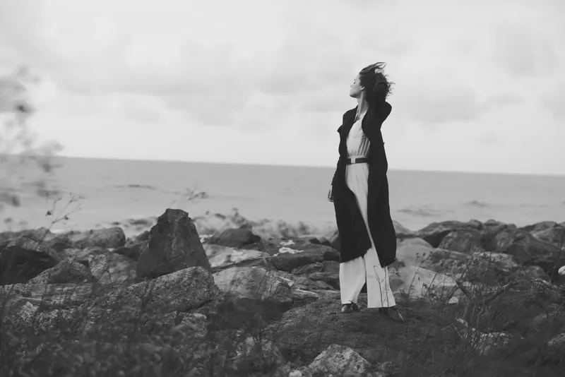 Woman stands on rugged rocks by the sea, facing the wind with her coat fluttering and dress flowing, capturing a moody outdoor landscape and a sense of freedom in monochrome