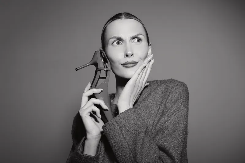 White woman with light skin holds antique telephone receiver, looking thoughtful, wearing sweater in studio with neutral background.
