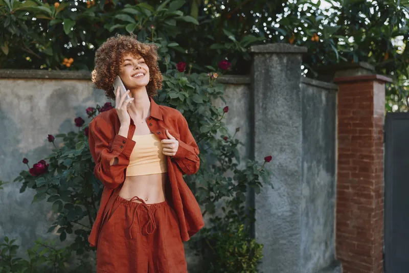 Young woman with curly hair outdoors wears a rust outfit while talking on the phone, smiling confidently, near a garden wall with leafy plants, soft natural light