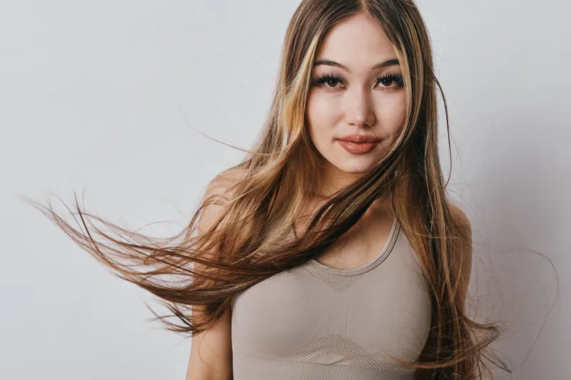 Young woman with long hair blowing and casual top posing in studio. Female portrait with natural makeup and confident expression on gray background with soft light.