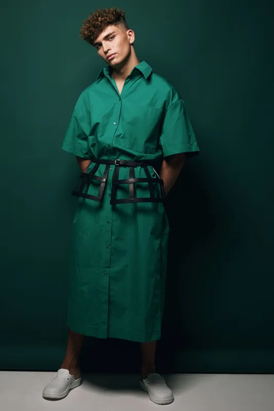 Young man posing in green oversized shirt dress with black belt accessory and white sneakers against dark green background in studio for fashion editorial.