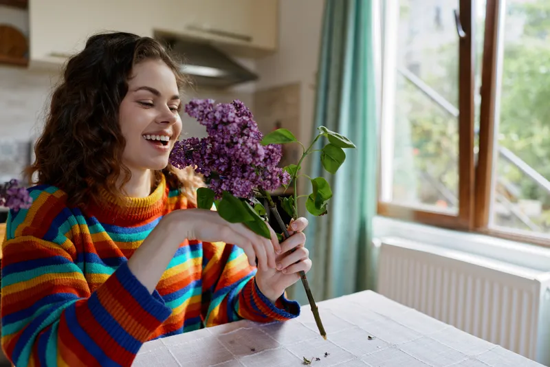 Woman with a colorful striped sweater smiles as she holds a lilac bouquet at a kitchen table, bright natural light, warm home mood, intimate lifestyle moment