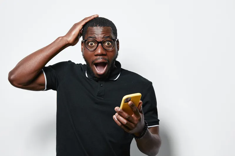 African American man with dark skin and glasses holding yellow smartphone with shocked expression and hand on head in casual black polo shirt on white background.