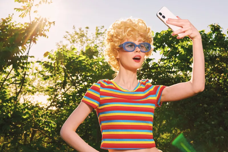 A woman with retro lifestyle poses for a selfie outdoors, striped shirt, curly blond hair, blue sunglasses, vintage film colors. Concept for nostalgia, casual joy, and summer moments.