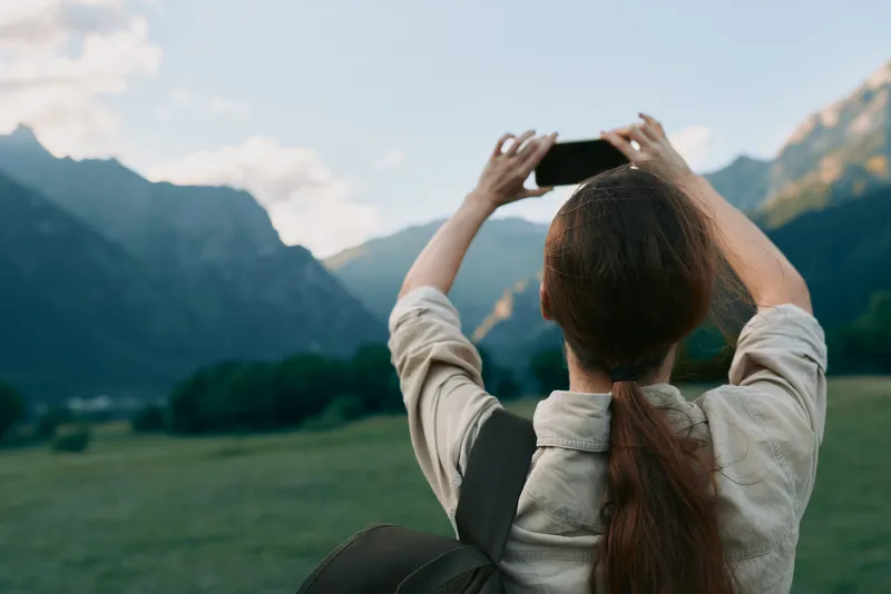 A female traveler photographed from behind uses a phone to capture a dramatic mountain landscape in a bright valley.