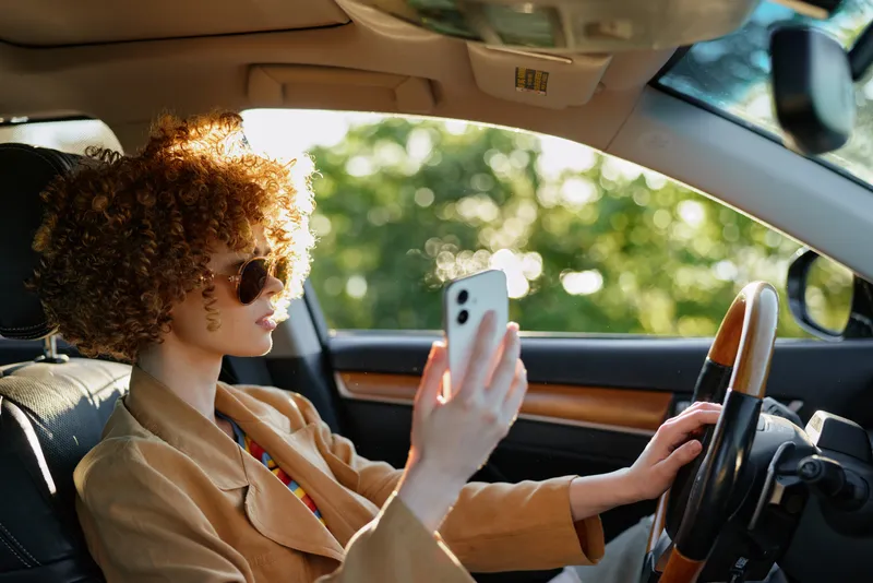 Bright candid scene of a woman driving a car, wearing sunglasses and using a smartphone, sunlit interior light with retro film color and warm tones, emphasizing lifestyle and casual style