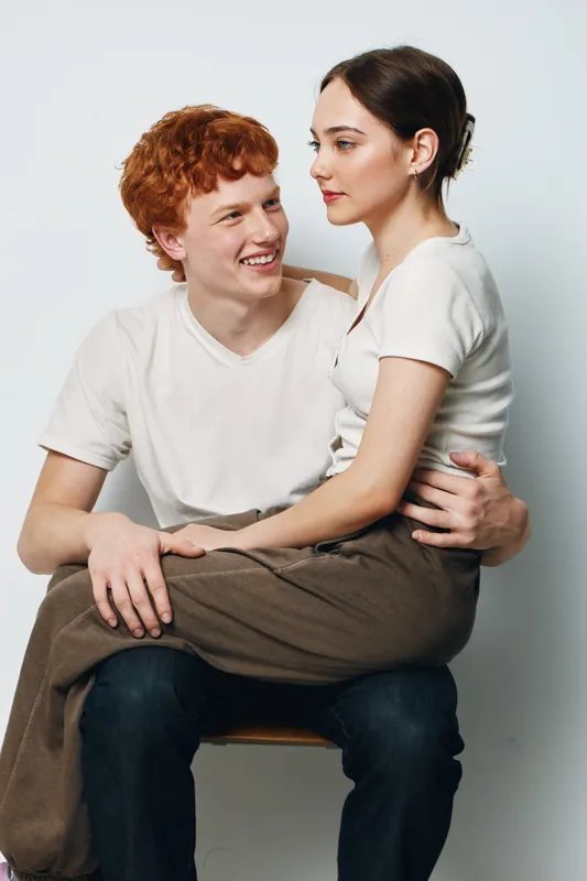 Young man with red hair smiling while holding woman in brown skirt sitting on his lap in white background studio portrait. Couple, love, affection and casual fashion.