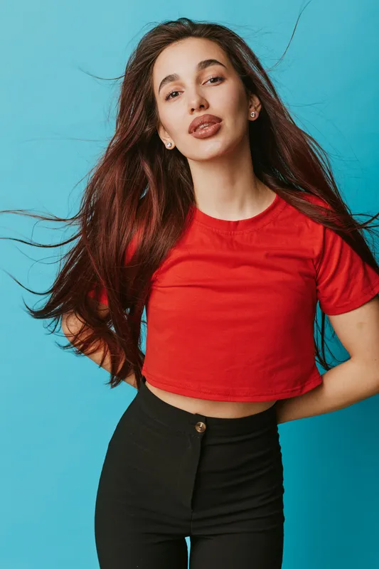 Young woman with long hair wearing red crop top and black pants posing against blue background. Casual style, confident expression, fashion portrait with natural makeup and earrings.