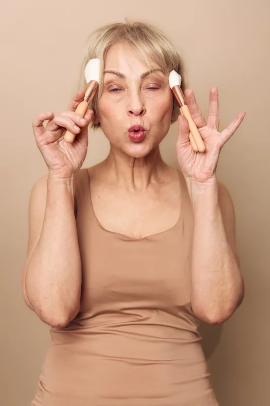Mature woman with short blonde hair applies makeup with brushes on beige background. Concept of beauty routine, skincare, natural look, and self-care for older women in studio lighting.