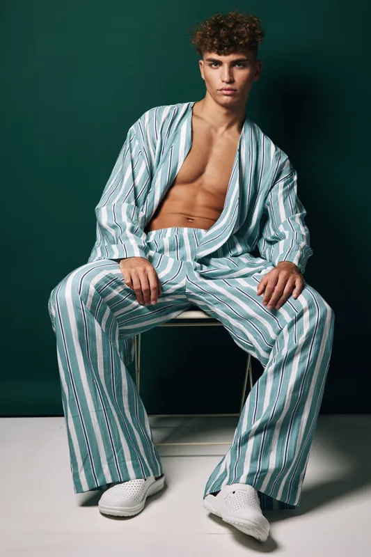 Young man with curly hair wearing casual striped shirt and pants sitting on chair against green background. Masculine, relaxed posture with open stylish top and white sneakers.