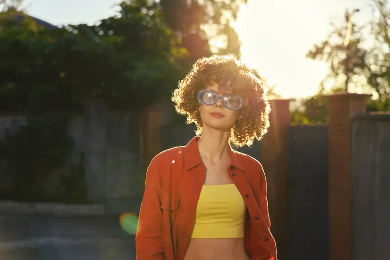 A stylish woman stands outdoors in warm sunlight, wearing a yellow crop top and red jacket, with sunglasses. The scene conveys casual confidence and sunny lifestyle