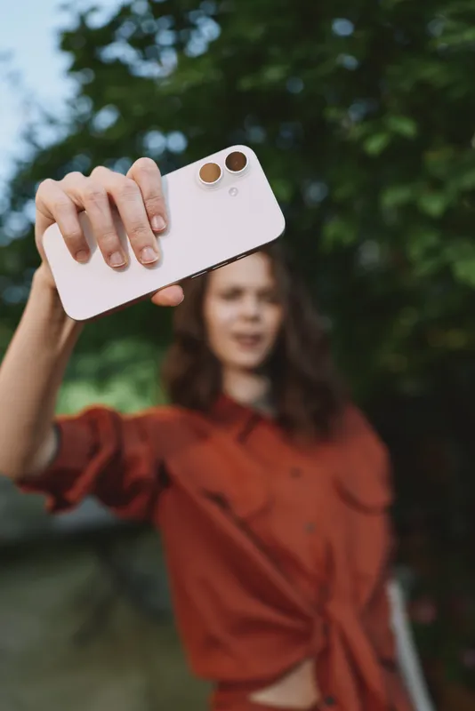 Cheerful woman takes a selfie outdoors with a smartphone, wearing casual clothes, sunlight and nature softly blurred in the background, a relaxed lifestyle moment