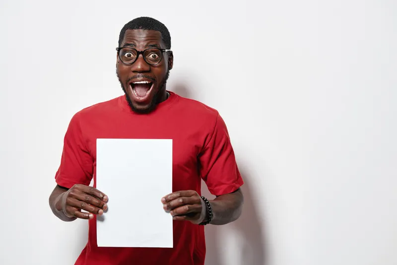 African American man with dark skin and glasses wearing red shirt holding blank white paper with excited expression on white background for presentation or announcement.