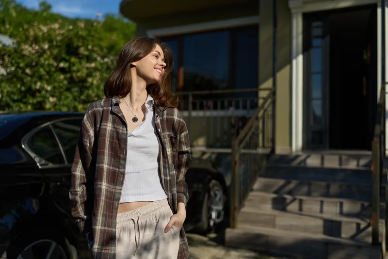 A woman in casual outdoor attire smiles in daylight, showcasing a relaxed lifestyle moment on a sunny street, with a confident fashion sense