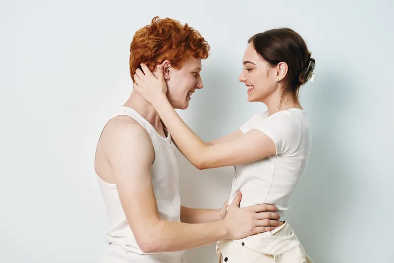 Young couple, man and woman embracing and smiling, intimate moment with affection and love. Casual clothing, standing face to face on white background with happiness and connection.
