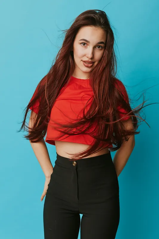 Young woman with long hair wearing red crop top and black pants posing with hands behind back on blue background. Casual fashion, confident, smiling and standing portrait.