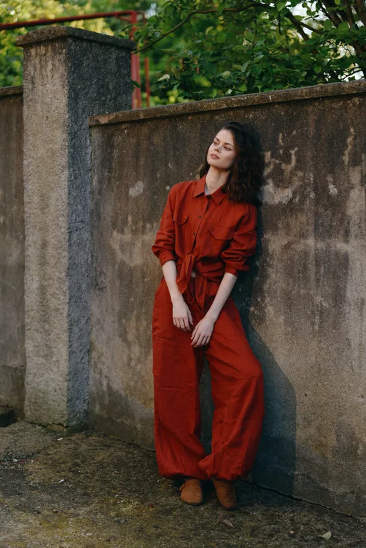 Woman in a red jumpsuit leans against a weathered wall outdoors, showcasing casual fashion, relaxed confident mood, natural light, and a simple backdrop with greenery, capturing everyday lifestyle