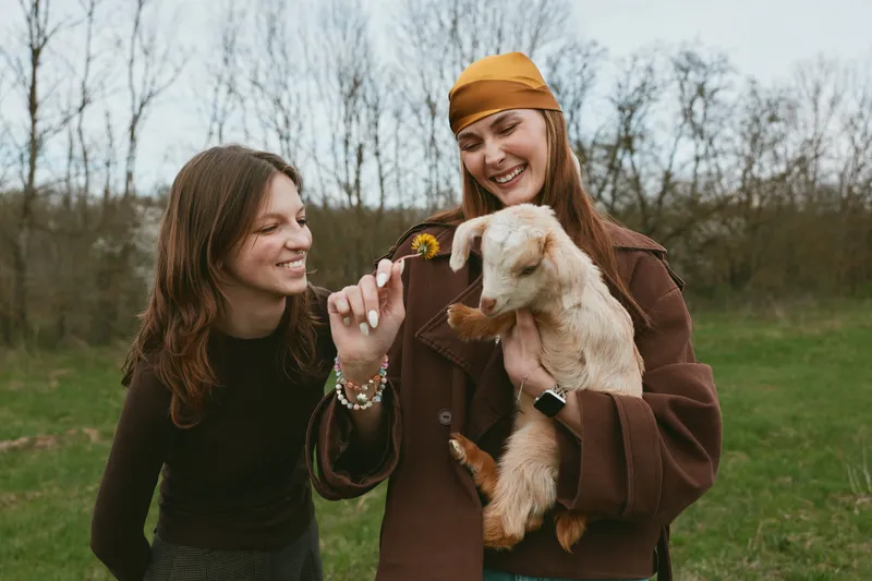 Two women outdoors share a gentle moment with a goat kid, highlighting friendship, affection, and lifestyle in a rural outdoor scene. Natural light, warm tones, casual outfits, and a tender animal