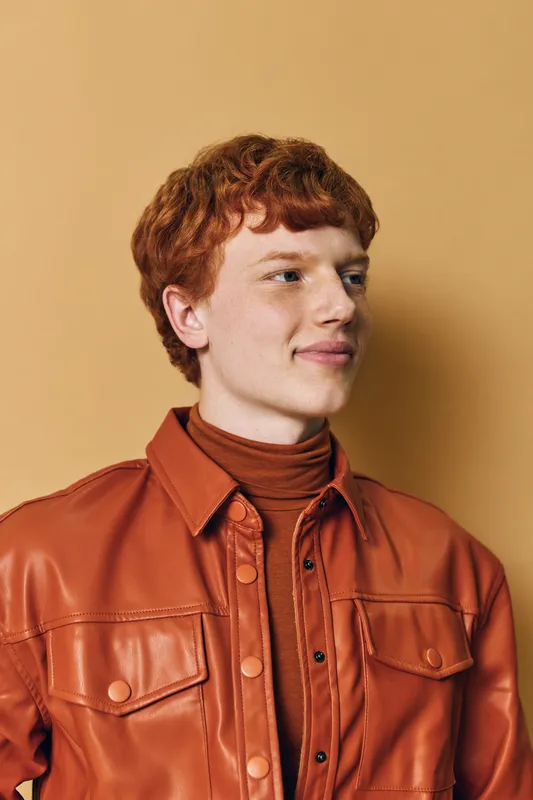 Young man with red hair wearing brown leather jacket and turtleneck standing against beige background looking sideways with slight smile and relaxed posture.