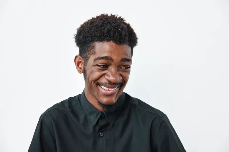 Young black man with dark skin and curly hair smiling and looking down wearing black shirt on white background. Portrait of happy African ethnicity male with casual style.