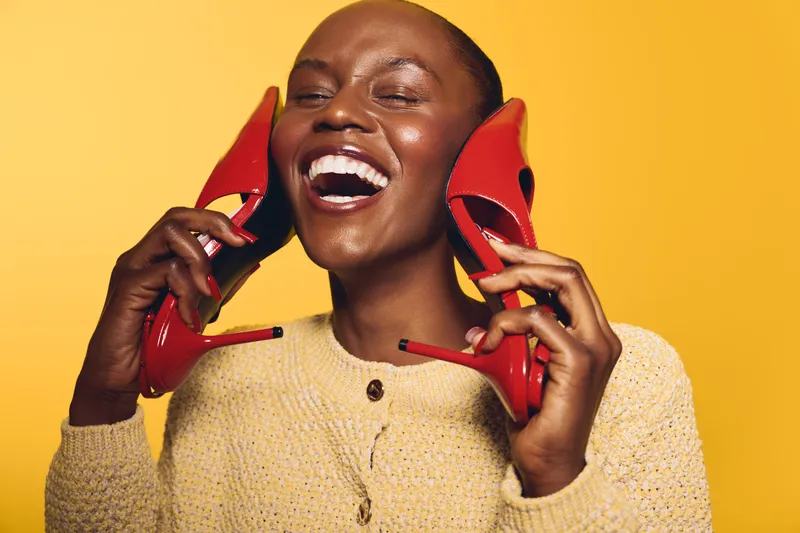 Happy African woman with dark skin laughs holding red high heels near her ears on yellow background. Concept for fashion, joy, lifestyle, and beauty with colorful style.